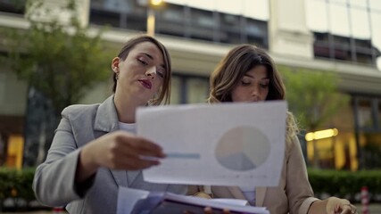 Professional business women engage in an important meeting, discussing reports while working together in an outdoor urban environment. They analyze financial data and strategic for their project.