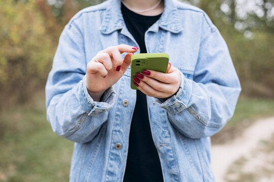 Female hands with red manicure holding green phone in denim jacket.