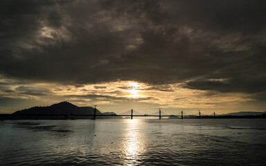 A scenic view of a cable-stayed bridge over the Brahmaputra River during sunset, with reflections shimmering on the water and dramatic clouds in the sky
