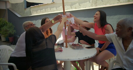 Diverse group of friends and family cheerfully raising cups in unison, toasting together at an...