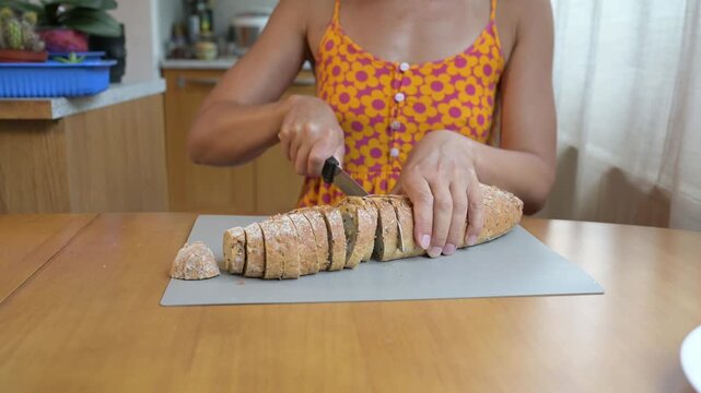 Close-up video of a woman slicing fresh multigrain bread with a knife on a cutting board at the kitchen table, preparing food at home