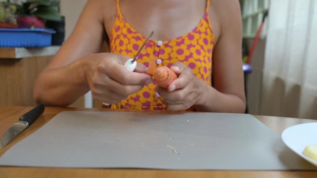 Close-up video of a woman peeling a fresh carrot with a knife over a plate at the kitchen table, preparing vegetables for cooking