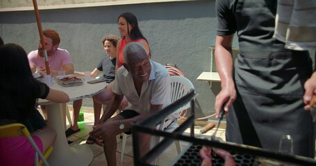 Man grilling food on barbecue with sausages and chicken skewers, diverse group of friends and family bonding at backyard summer celebration