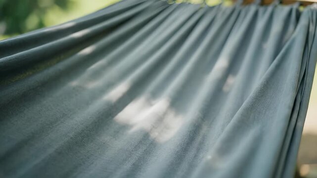 The gray hammock sways gently as light casts shadows on the gray fabric. The hammock provides a serene rest under the trees, with soft folds in the gray material.
