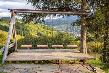 A rustic wooden swing set on a platform overlooks a scenic valley with mountains, forests, and a village in the distance on a sunny day.