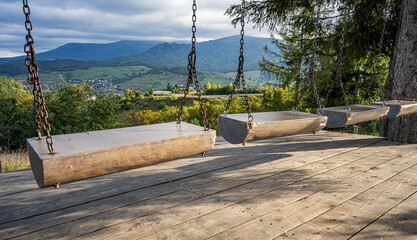 A rustic wooden swing set on a platform overlooks a scenic valley with mountains, forests, and a village in the distance on a sunny day.