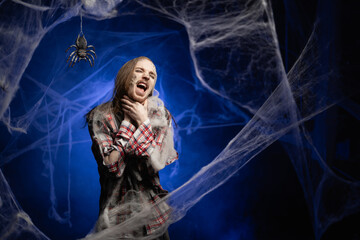 Creepy scary zombie man with ugly makeup shouting and suffocate because painful strangle health problem standing on spooky spider web background. Halloween. Horror film.