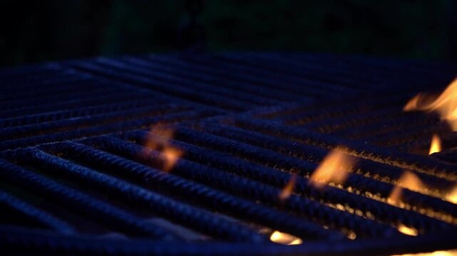 Flames rising through metal grill grates in a dark scene, capturing intense heat, fire motion, and dramatic barbecue atmosphere with strong contrast and texture.