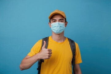 Young man wearing yellow casual t-shirt, face mask and cap, showing thumbs up with backpack on blue background, representing travel and outdoor adventure concepts.