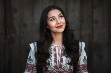 Fototapeta premium Portrait of a young woman with long wavy dark hair and red lips standing in front of a wooden barn in a traditional embroidered folk blouse looking upwards with a thoughtful expression