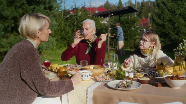 Side view shot of middle-aged Caucasian mother talking to teenage daughter sitting around festive table on sunny autumn day, spending family time together in countryside