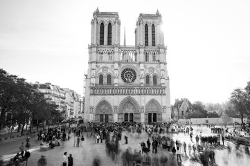 A lively scene unfolds as people gather near Notre-Dame-de-Paris at sunset. The historic cathedral's architecture is beautifully highlighted in black and white, capturing the moment.