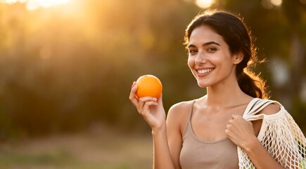 Happy beautiful woman holding a fresh orange and a reusable mesh bag enjoying the golden hour sunlight. Healthy eating and eco-friendly lifestyle concept