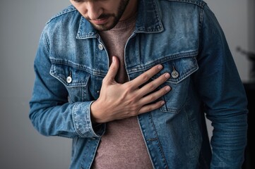 Young man in denim jacket at home holding chest indicating discomfort or pain expression of health issue or feeling unwell indoors setting