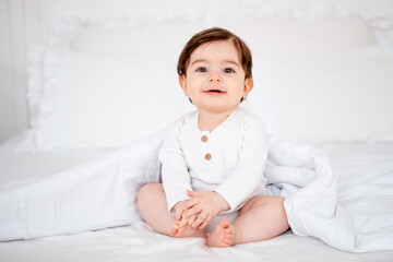 A baby under a blanket on a white bed at home, a place for text, a cute smiling little child getting ready for bed