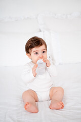baby with a bottle of milk or water on a white bed at home, baby food feeding concept, place for text, cute smiling little baby