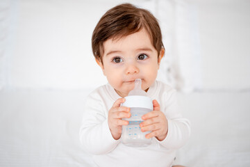 baby with a bottle of milk or water on a white bed at home, baby food feeding concept, place for text, cute smiling little baby