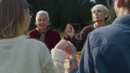 Zoom out shot of delighted middle-aged Caucasian mother saying grace holding hands with family members while sitting at table outdoors on warm autumn day