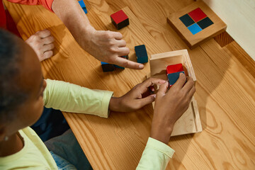 Child assembling wooden cubes with teacher guidance at desk. Concept of hands-on learning, psychology, healthcare, EdTech, medical marketing, and publishing for education.