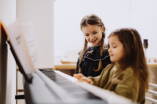 Two young girls playing a piano together, one smiling and looking at the other. The setting is bright and modern, with sheet music visible on the stand.