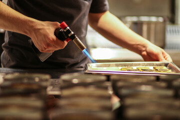 A chef using a culinary torch to finish or caramelize small portions of food on a tray in a...