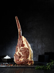 A large tomahawk steak stands upright on a dark stone surface, highlighted by its impressive marbling. Nearby, a small bowl holds salt, while fresh herbs add a touch of color. © Vitte Yevhen