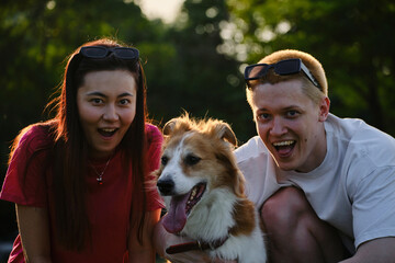 Young couple making funny faces with their dog in the park. Playful outdoor lifestyle with pets.