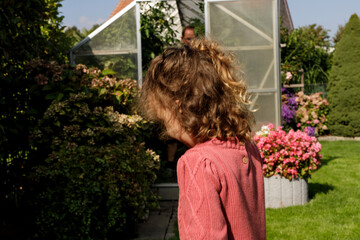 A young child in a pink sweater explores a vibrant garden with flowers, lush greenery, and a greenhouse.  Captured on a sunny day, evoking a sen © vegefox.com