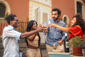 Happy diverse friends toasting with wine at a summer celebration. Multicultural group laughing and enjoying a party with togetherness outdoors in a rustic courtyard, sharing candid moment of pure joy.
