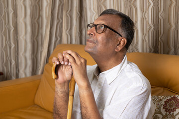 Indian Lonely old man holding wooden stick at home. Unhappy old man sitting on sofa at home,...