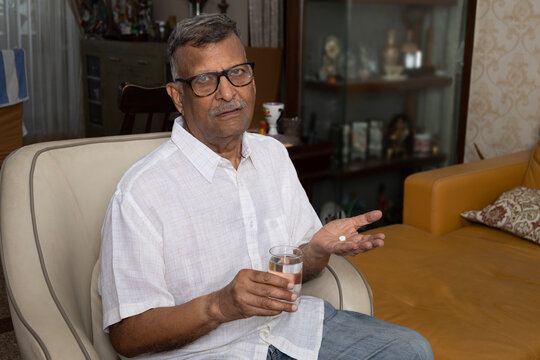 Indian senior unhealthy man taking a pills and a glass of water while sitting at home - Powered by Adobe