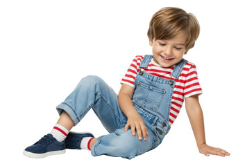 Smiling little boy in denim overalls and striped shirt isolated on transparent background, happy child sitting and looking down