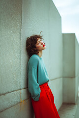 A woman in a blue sweater and red pants is leaning against a wall