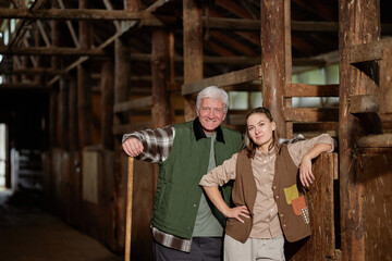 Caucasian senior man and Caucasian young adult woman standing together in rustic barn, both smiling...