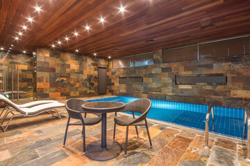 The elegant design of the indoor swimming pool features stone walls and a warm-colored wooden ceiling. There are comfortable sun loungers and a small table for relaxing.