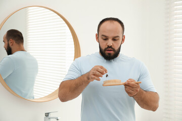 Baldness problem. Worried man taking his lost hair from brush near mirror in bathroom