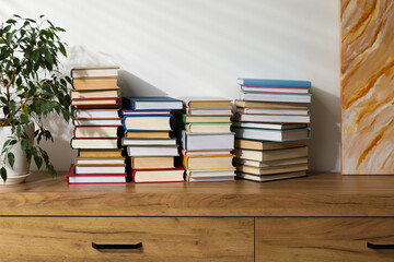 Many stacked books on wooden chest of drawers and plant indoors