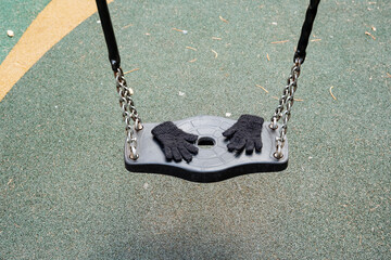 A swinging playground swing, completely empty, sways gently in the calm breeze, with a pair of black gloves resting on its seat, stirring feelings of nostalgia and leisure in the deserted park area