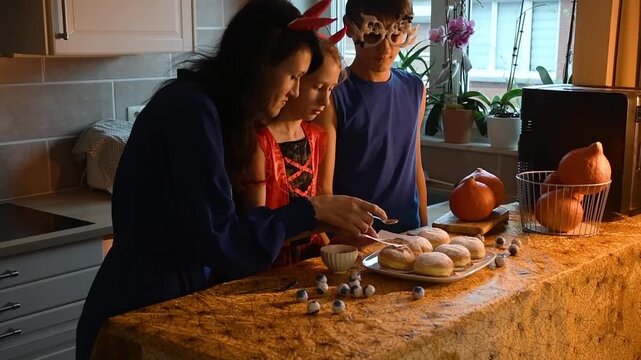 Happy mother and children in festive costumes decorate homemade donuts with cocoa powder, draw skull templates to create Halloween decor, eye-shaped candies on table,spooky and fun holiday tradition