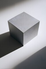 A grey cube sits on a white surface, illuminated by a light source casting a prominent shadow. The composition highlights geometric forms and spatial relationships.