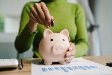 Woman putting coin into pink piggy bank on desk, symbolizing saving, investment, financial planning, retirement, and smart money management.