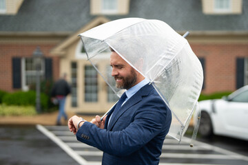 Businessman waits under the rain holding an umbrella. A confident man checks his watch while standing on a wet street. Businessman checks the watch. Man watching the clock. A punctual business man.