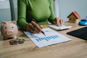 A person analyzing financial charts with a calculator, piggy bank, house model, and car model,...