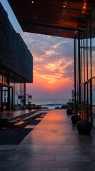 A modern building's walkway frames a vibrant sunset over the ocean, featuring glass walls, stone textures, and potted plants. The sky transitions from orange to blue.