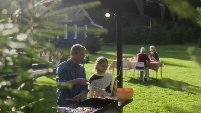 Teenage Caucasian girl helping his father to grill vegetables spending warm autumn weekend in cottage house with family sitting at table outdoors in background