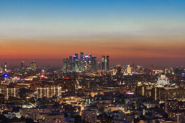 As the sun sets, the city skyline glimmers with bright lights. The scene captures a mix of modern structures and historic architecture against a gradient sky.