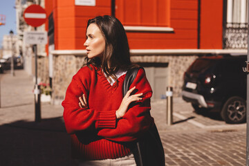 Confident young woman wear red sweater posing with crossed arms against red building standing in warm sunlight. Natural light fashion portrait with stylish attitude and relaxed posture.