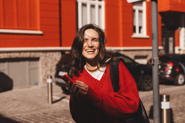 Brunette long hair woman holding mobile phone, look at camera, laughing and smiling, good news, scrolling media, she walking outdoor on the street town. Woman wear red sweater, black bag.