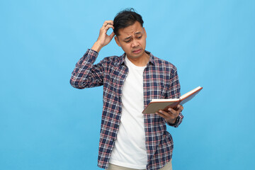 A confused young man scratching his head, planning his schedule, reading a diary or journal he is holding, standing against a blue background.