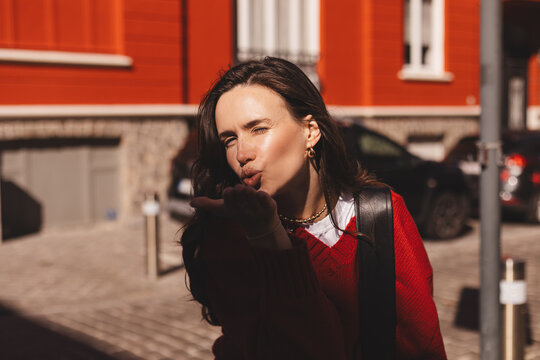 Portrait close up brunette long hair woman send blow air kiss, wear red sweater. Woman outdoors blowing kiss on city street evokes warm expression featuring a casual outfit. 